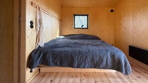 Une petite pièce chaleureuse, avec un décor intérieur en bois, un lit simple recouvert d’une couverture sombre, une petite fenêtre donnant sur un paysage agréable et un plancher en bois.