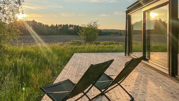 Une terrasse extérieure moderne, dotée de deux chaises pliantes noires, d’un plancher en bois et d’un mur en verre offrant une vue sur un champ et des arbres.