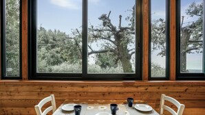 Une table à manger, avec des assiettes, des tasses et des couverts pour quatre personnes, est placée devant une fenêtre donnant sur des arbres et un ciel dégagé.