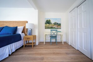 A bedroom with a bed, bedside table, lamp, desk, chair, and a painting on the wall.