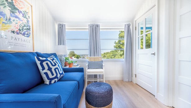 A bright room with a blue sofa, a wicker ottoman, a white chair, and a framed artwork on the wall. - Private Cottage at Marina Bay (Waterford)