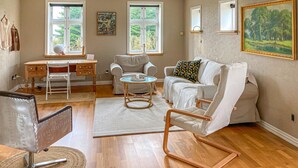 A spacious living room with a wooden floor, a white sofa, a white armchair, a small round table, and a desk with a chair. There are two windows with a view of greenery outside. - 3 bedroom cozy home in Havndal (Havndal)