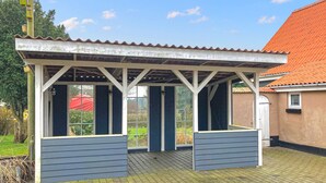 A covered outdoor seating area with a roof and windows, surrounded by a garden and a building with a red-tiled roof. - 3 bedroom cozy home in Havndal (Havndal)