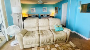 Ein Zimmer mit einem beigefarbenen Sofa, einem Standventilator und einem Deckenventilator. Zwei Nachttische mit Lampen sind vorhanden.