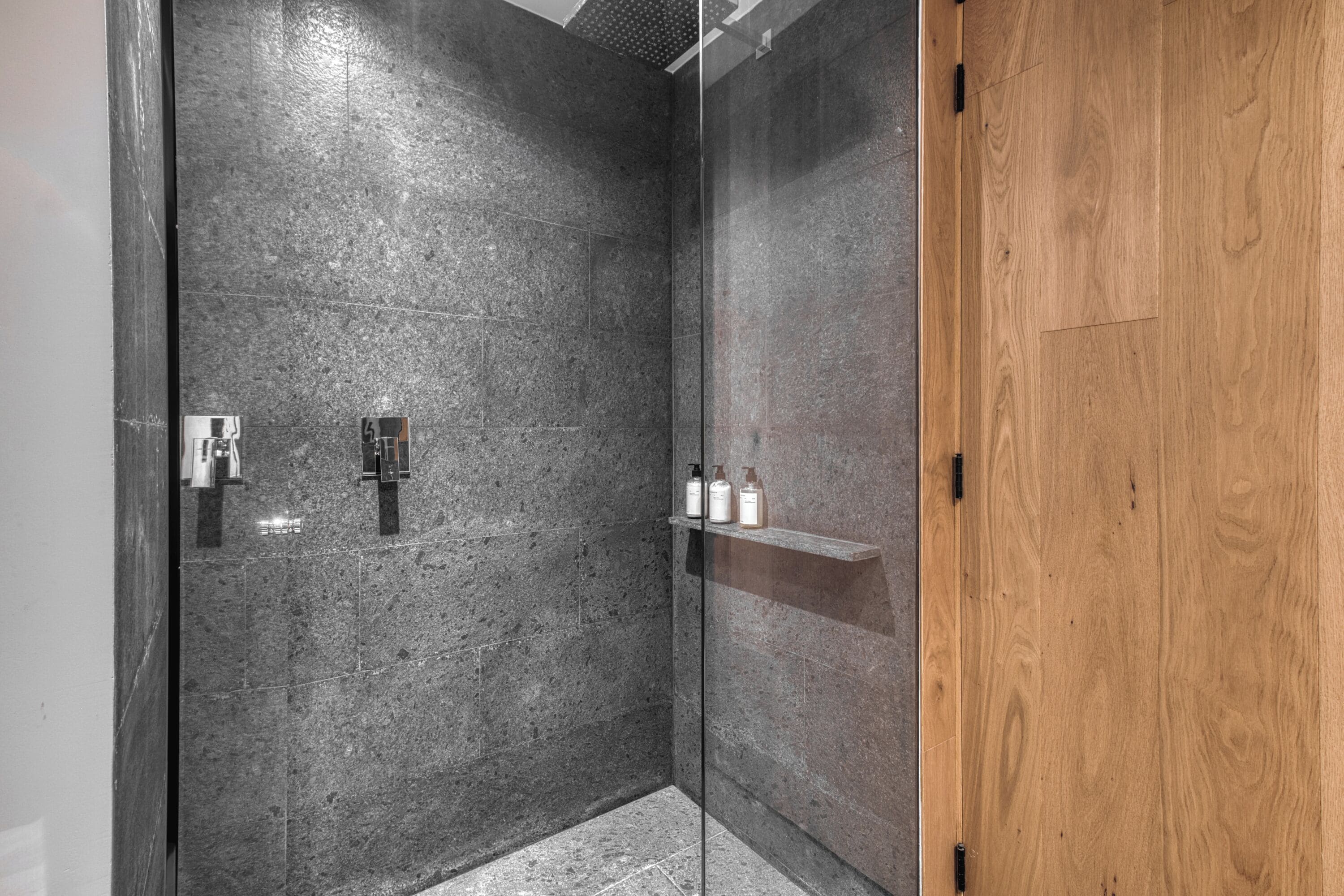 Un área de ducha moderna con cabina de vidrio, puerta de madera y repisa con artículos de aseo.