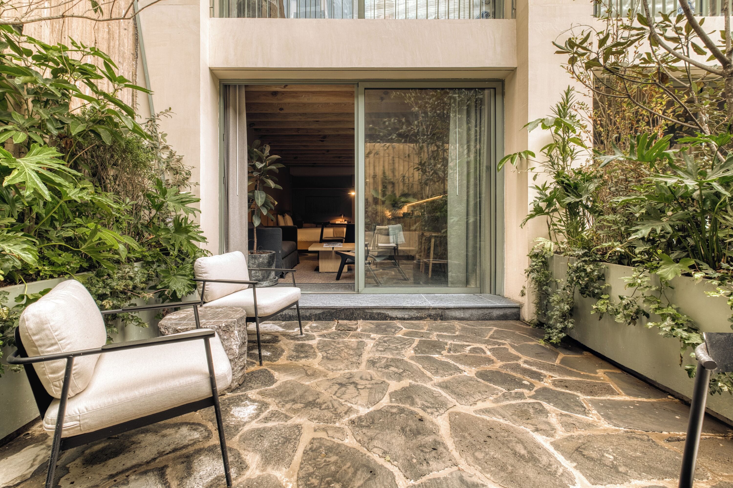 Un patio de piedra con mobiliario exterior, una puerta corrediza de vidrio que da a un espacio interior y abundante vegetación.