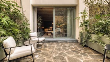 Un patio de piedra con mobiliario exterior, una puerta corrediza de vidrio que da a un espacio interior y abundante vegetación.