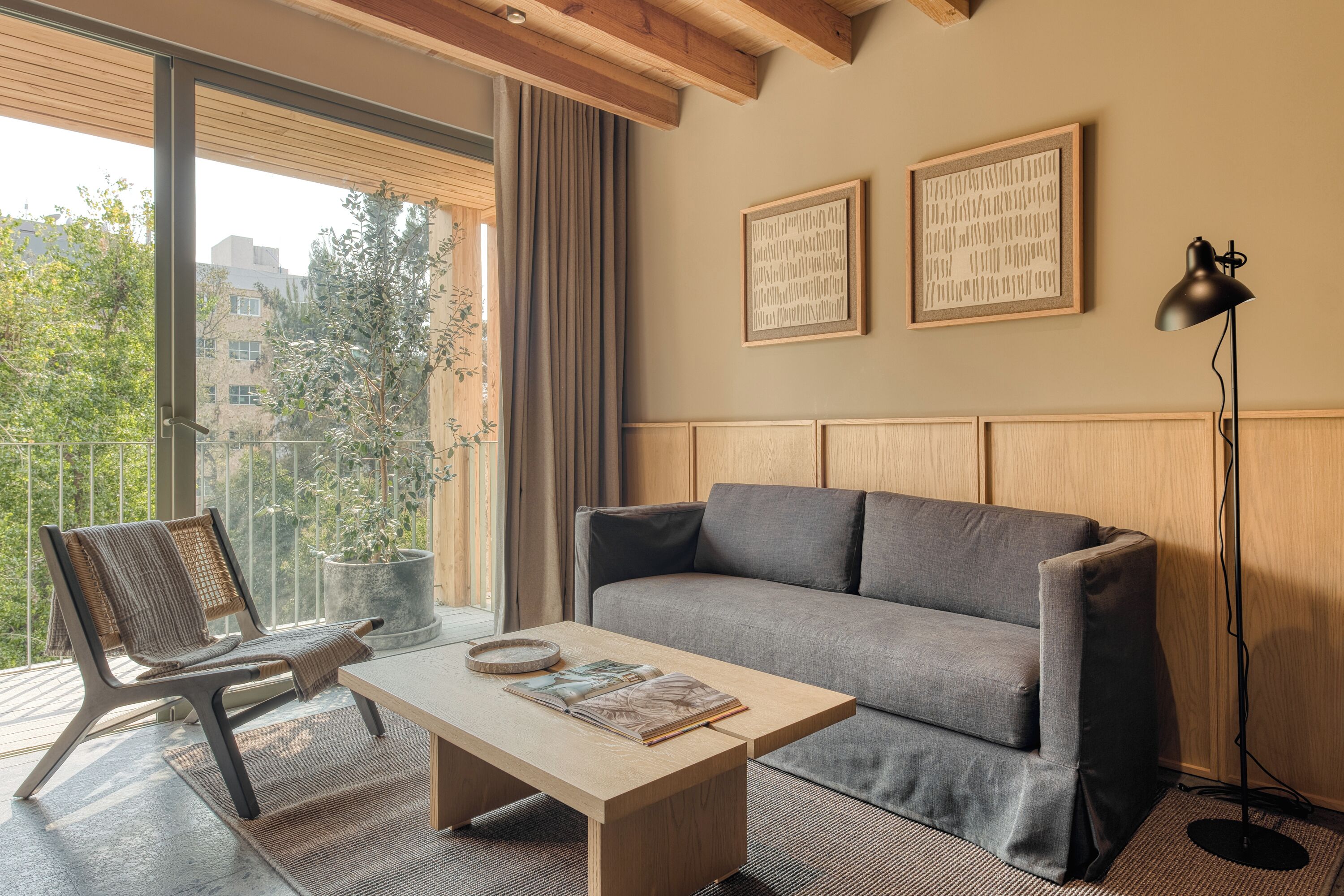 Un salon moderne avec un canapé gris, une table basse en bois et un fauteuil en osier. Deux œuvres d’art encadrées ornent le mur, et une lampe sur pied est placée dans un coin.
