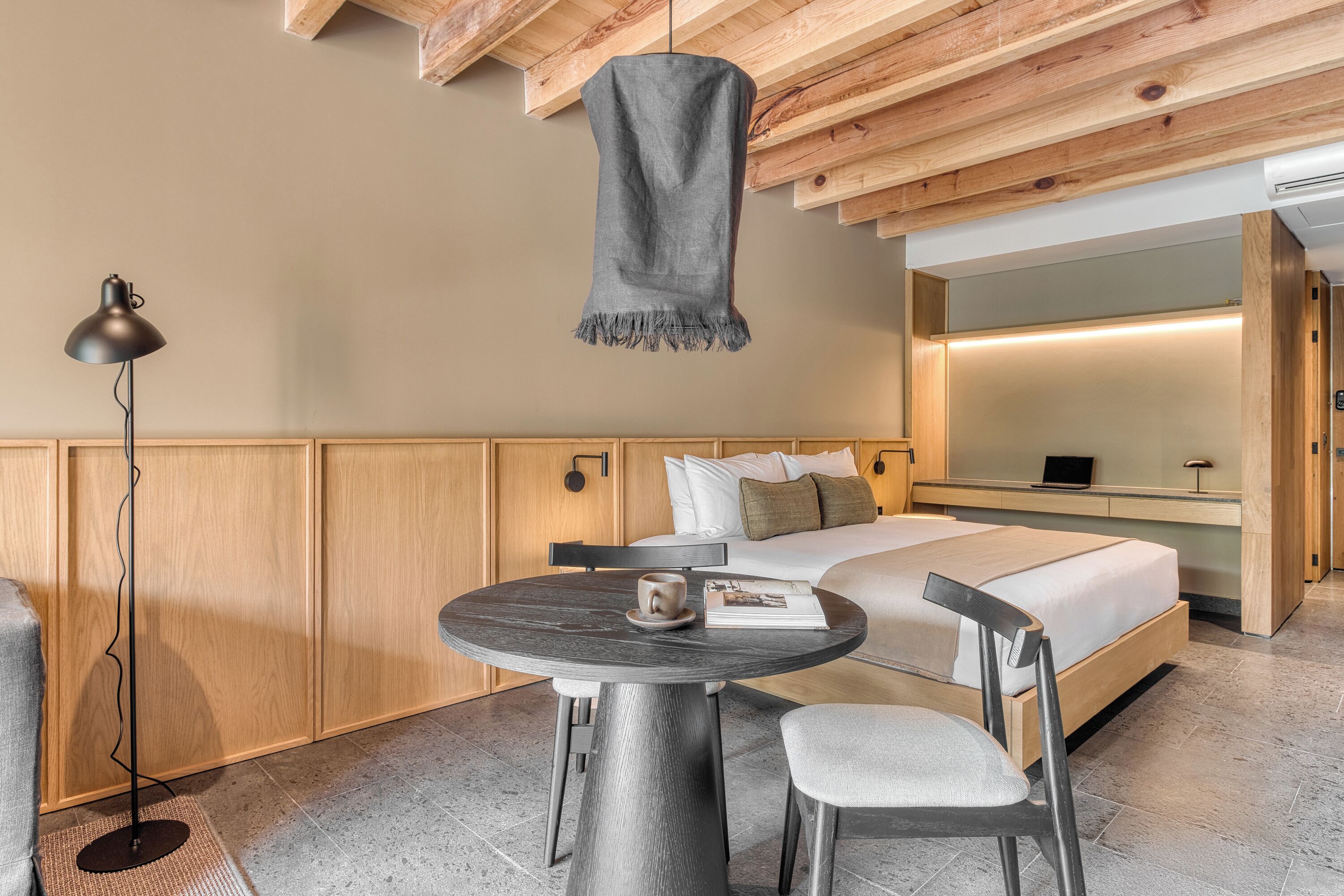 A modern hotel room with a wooden ceiling, a round table with a lamp, a bed with a white mattress, and a bedside table with a laptop.