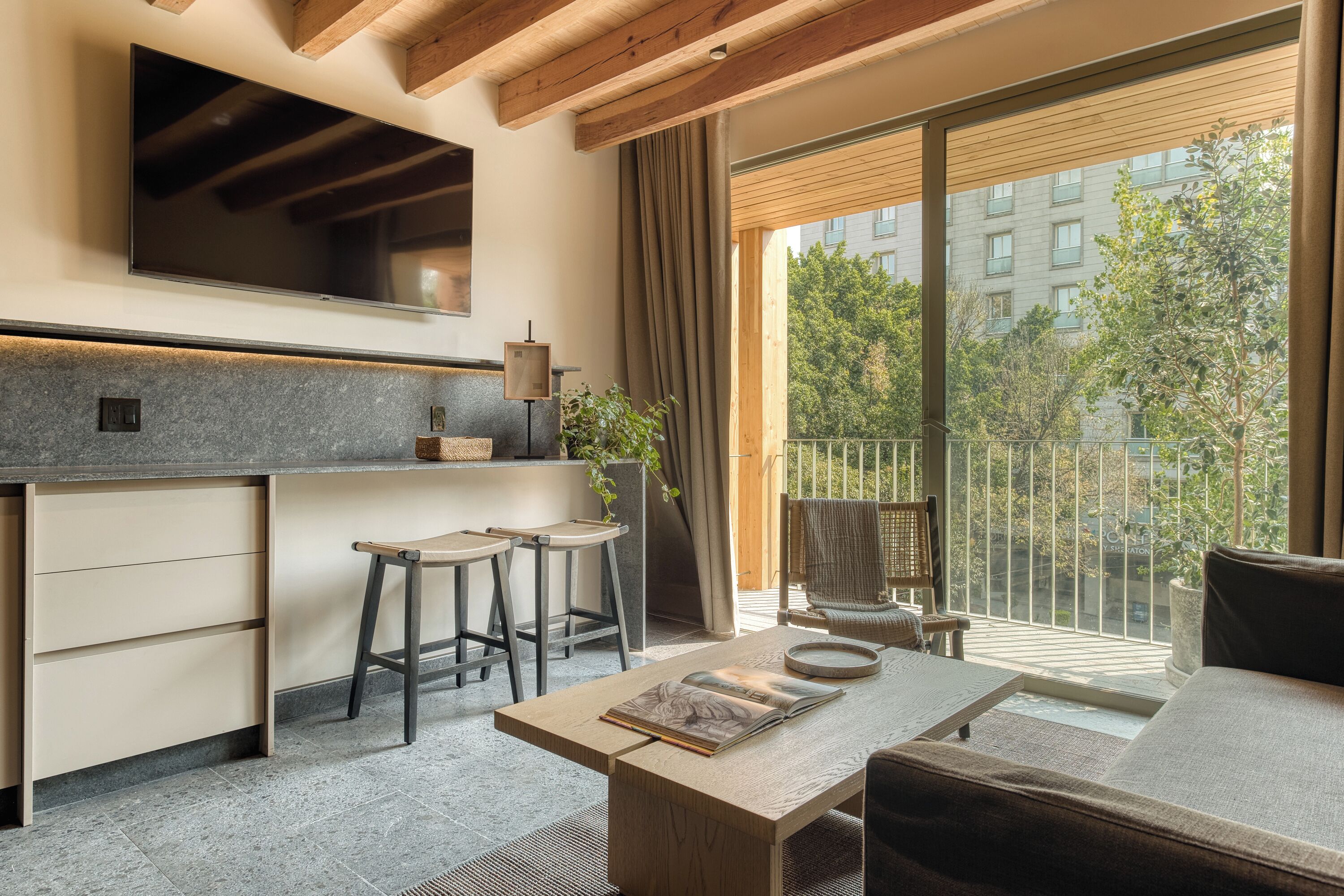 Eine moderne Küche mit großem Fenster, eine Essecke mit Sofa und Couchtisch, und ein Balkon mit Blick auf Bäume und Gebäude.