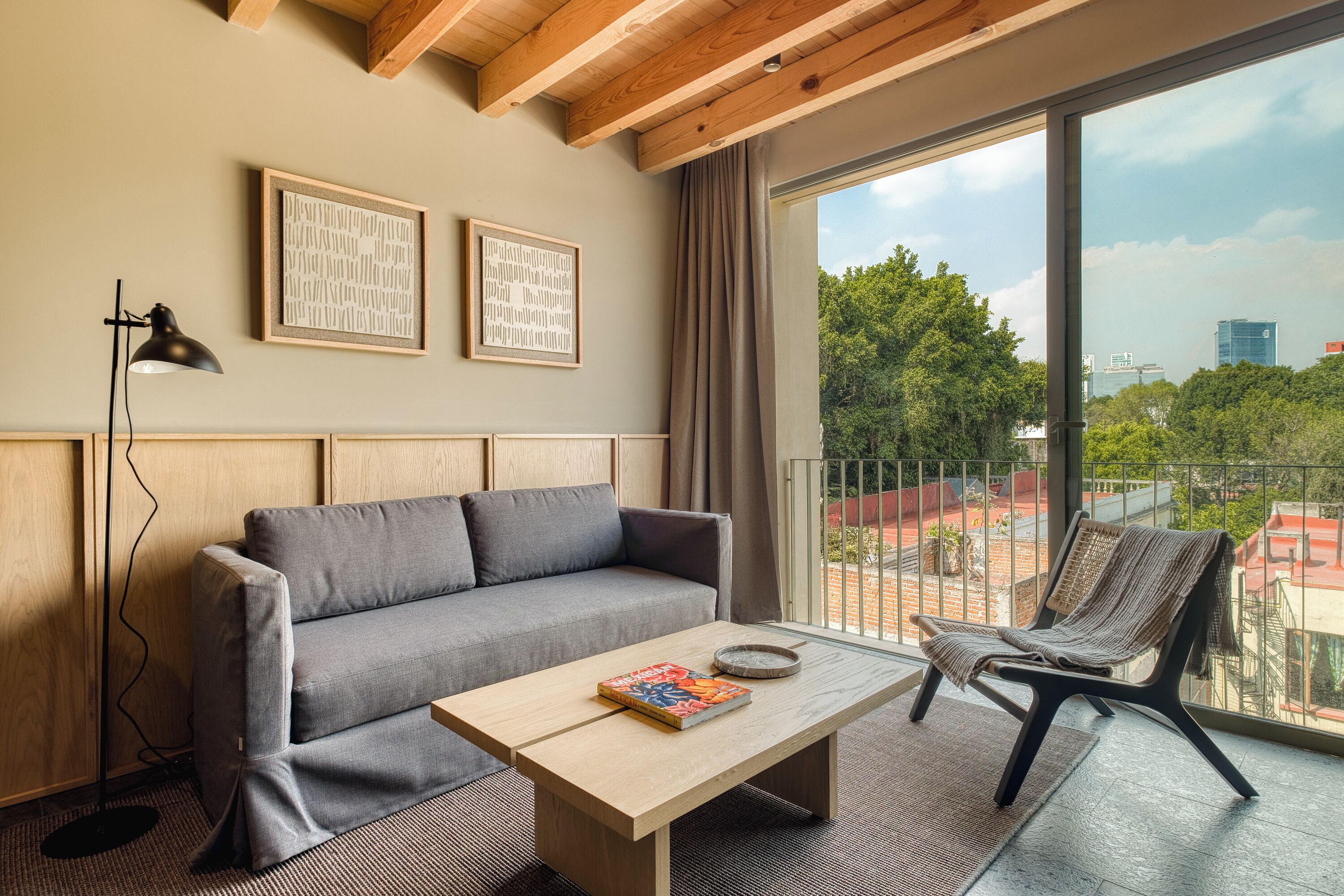 A modern living room with a grey sofa, a wooden coffee table with a book, and a chair. There are two framed artworks on the wall and a large window with a view of trees and buildings.