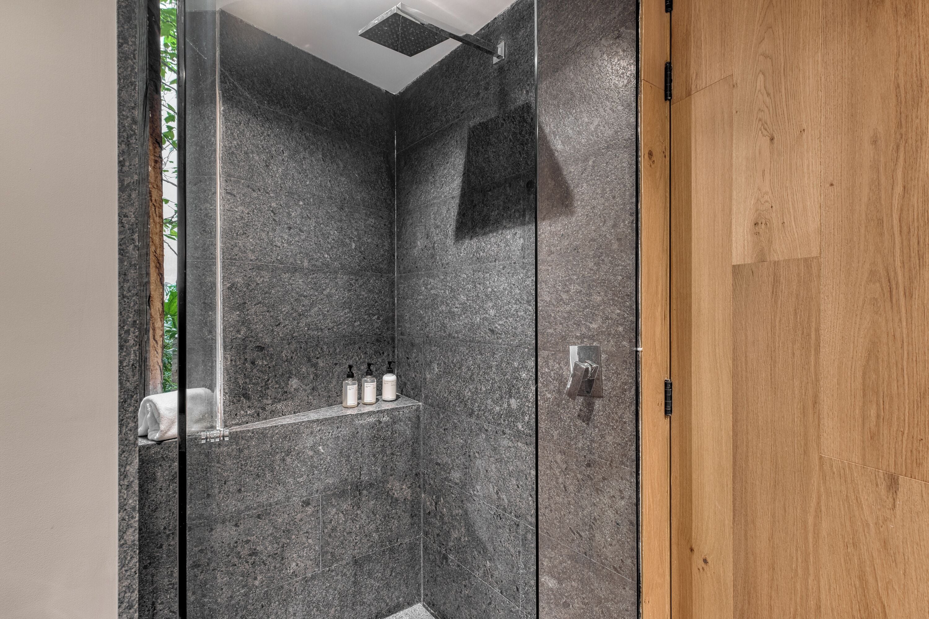 A modern shower with a glass door, a wooden door frame, and a shelf with toiletries.