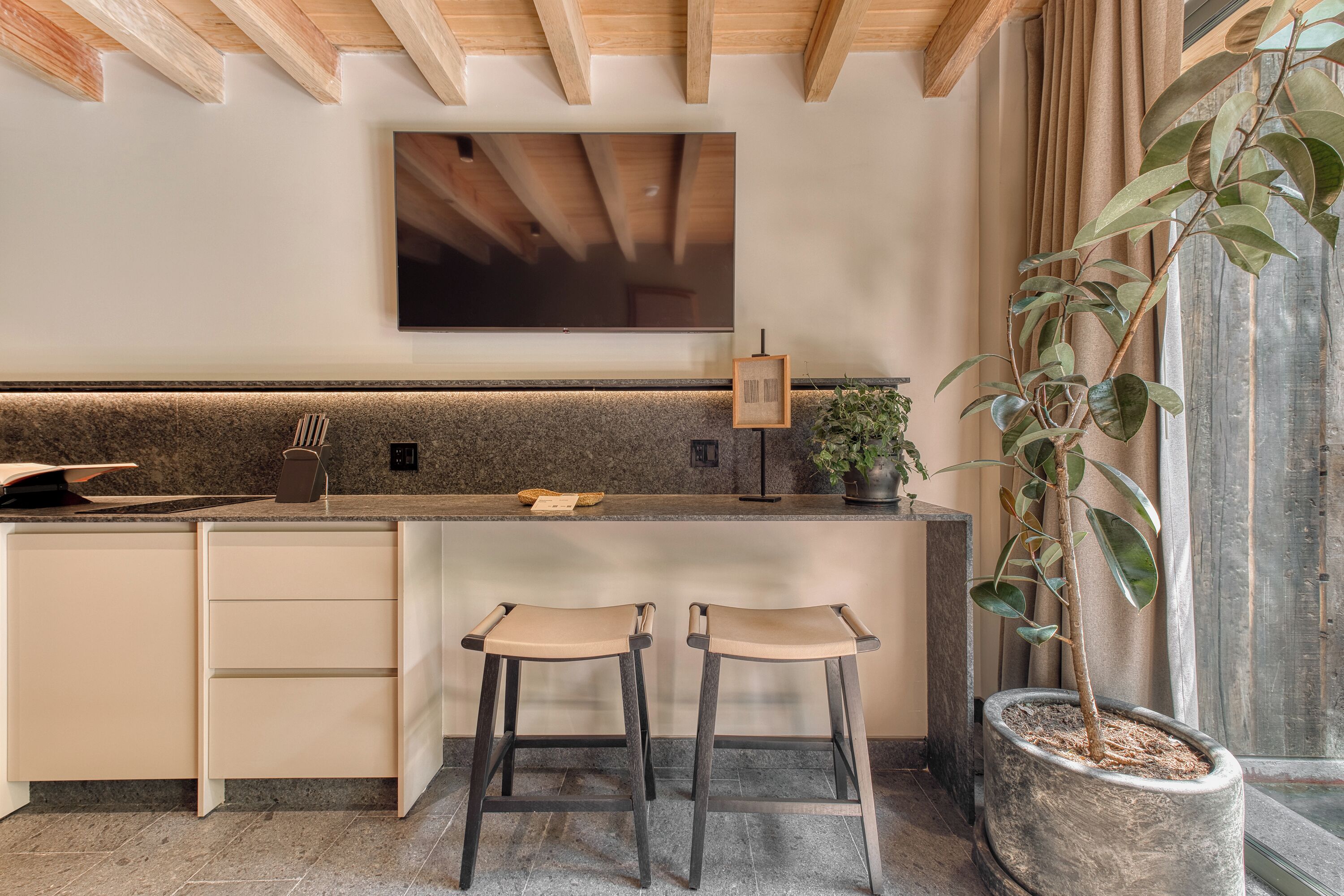 Cocina moderna con un televisor grande, dos taburetes de bar y una planta en maceta.