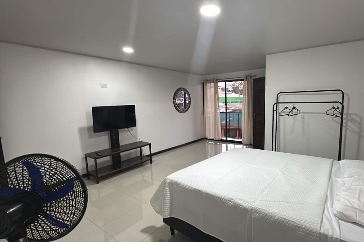 Un dormitorio con una cama, un televisor sobre un mueble, un ventilador y una ventana con cortinas.