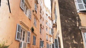 A narrow street with buildings on both sides, featuring arched doorways and shutters, and a sign indicating a location.
