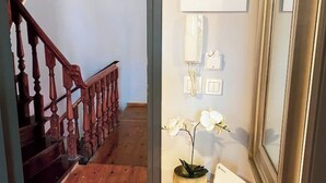 A hallway with a wooden staircase, a small table with a potted orchid, and a wall-mounted light switch. - Downtown Oasis (Kerkira)