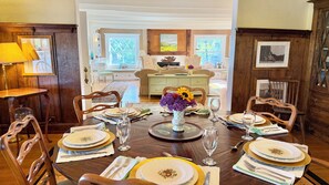 A dining room with a wooden table set for six, surrounded by chairs, with a view of a kitchen and living area in the background.