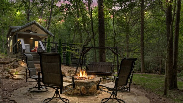 A stone fire pit with two chairs, a wooden swing set, and a cabin in a forest. - Creekside Tiny house in fantastic Monteagle! Quick hike to waterfall!  (Monteagle)