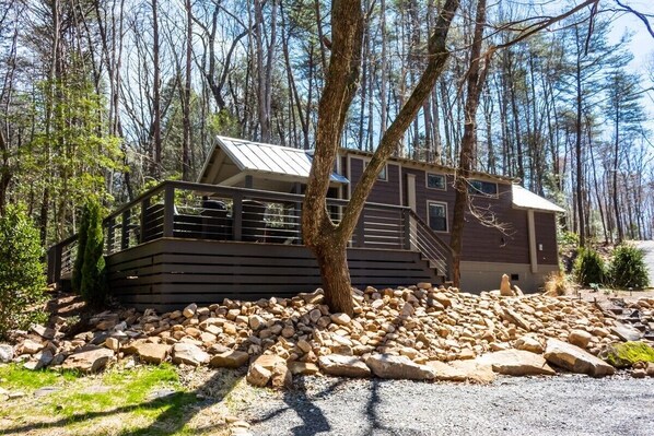 A cabin with a deck, surrounded by trees and a rocky ground.
