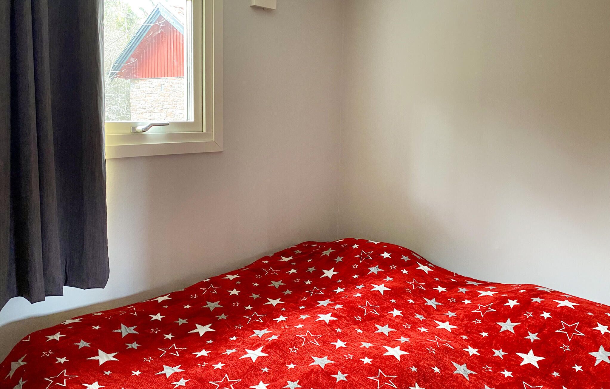 A room with a bed covered in a red blanket with white stars, a window with a view of a red building, and a white wall.