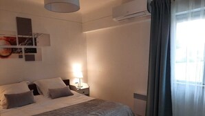 A bedroom with a bed, bedside lamp, air conditioning unit, and a window with curtains.