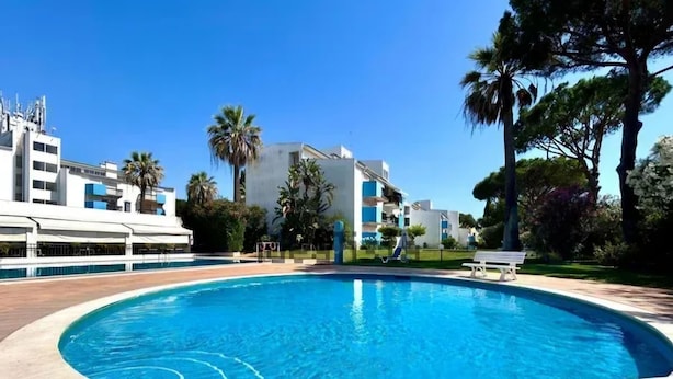 A swimming pool surrounded by a paved deck, with modern buildings and palm trees in the background.