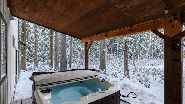 Un jacuzzi bajo una pérgola de madera con vista a un bosque nevado.