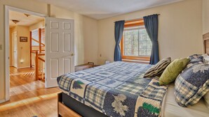 Un dormitorio con cama, ventana con cortinas, puerta que da a un pasillo y una escalera.