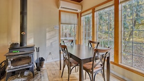 Un comedor con mesa y sillas, una estufa de leña y grandes ventanales con vistas a árboles.