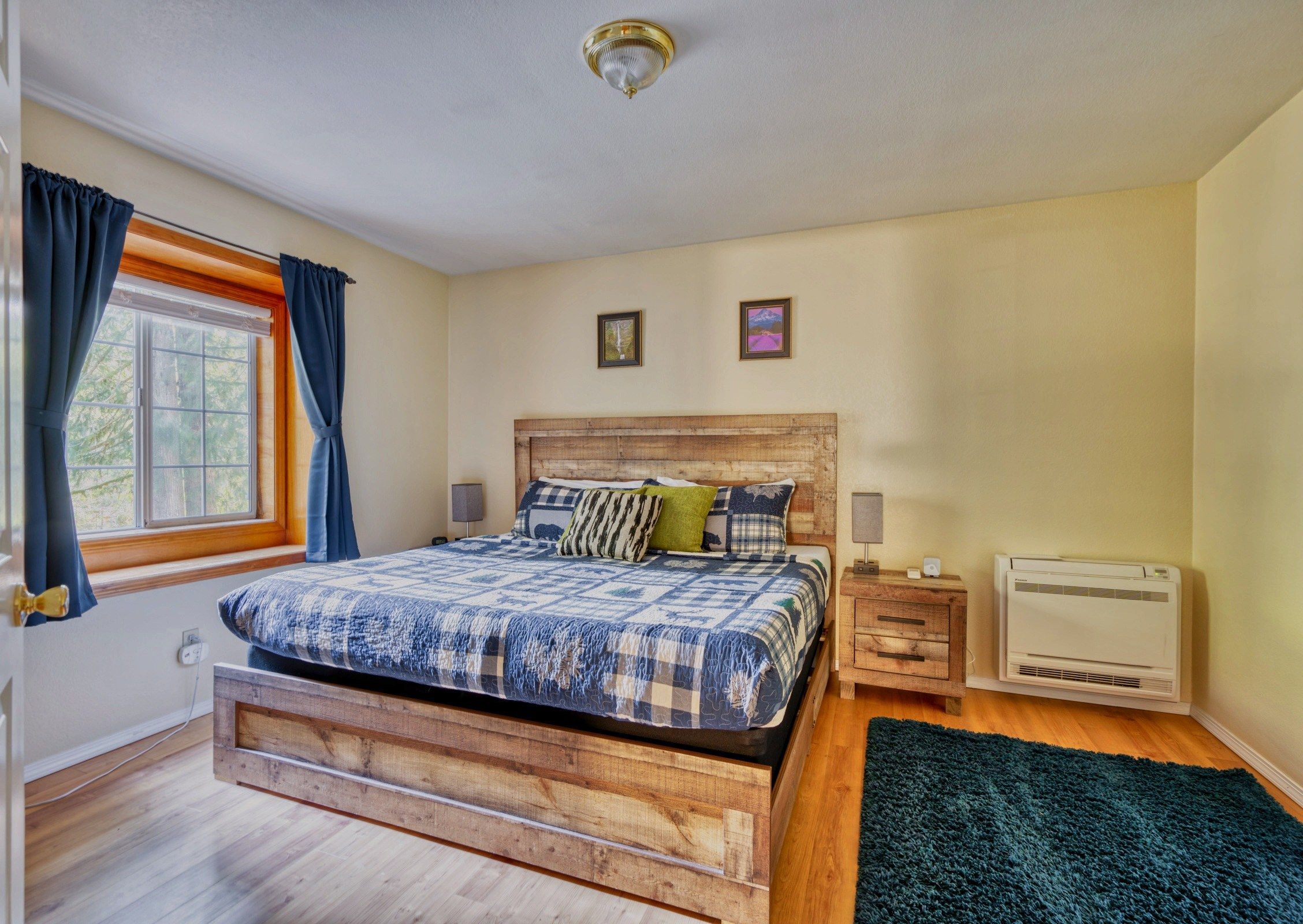 A bedroom with a wooden bed, a window with curtains, a wall-mounted air conditioner, and two framed pictures.