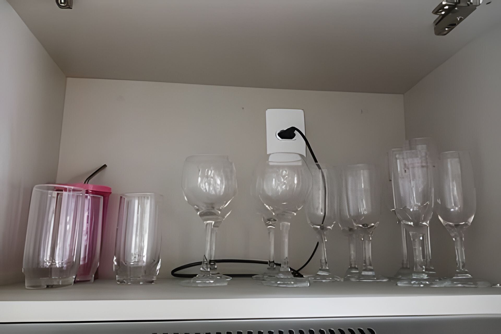 Una estantería con varios vasos y un dispositivo blanco, posiblemente un cargador, en una habitación de paredes blancas.