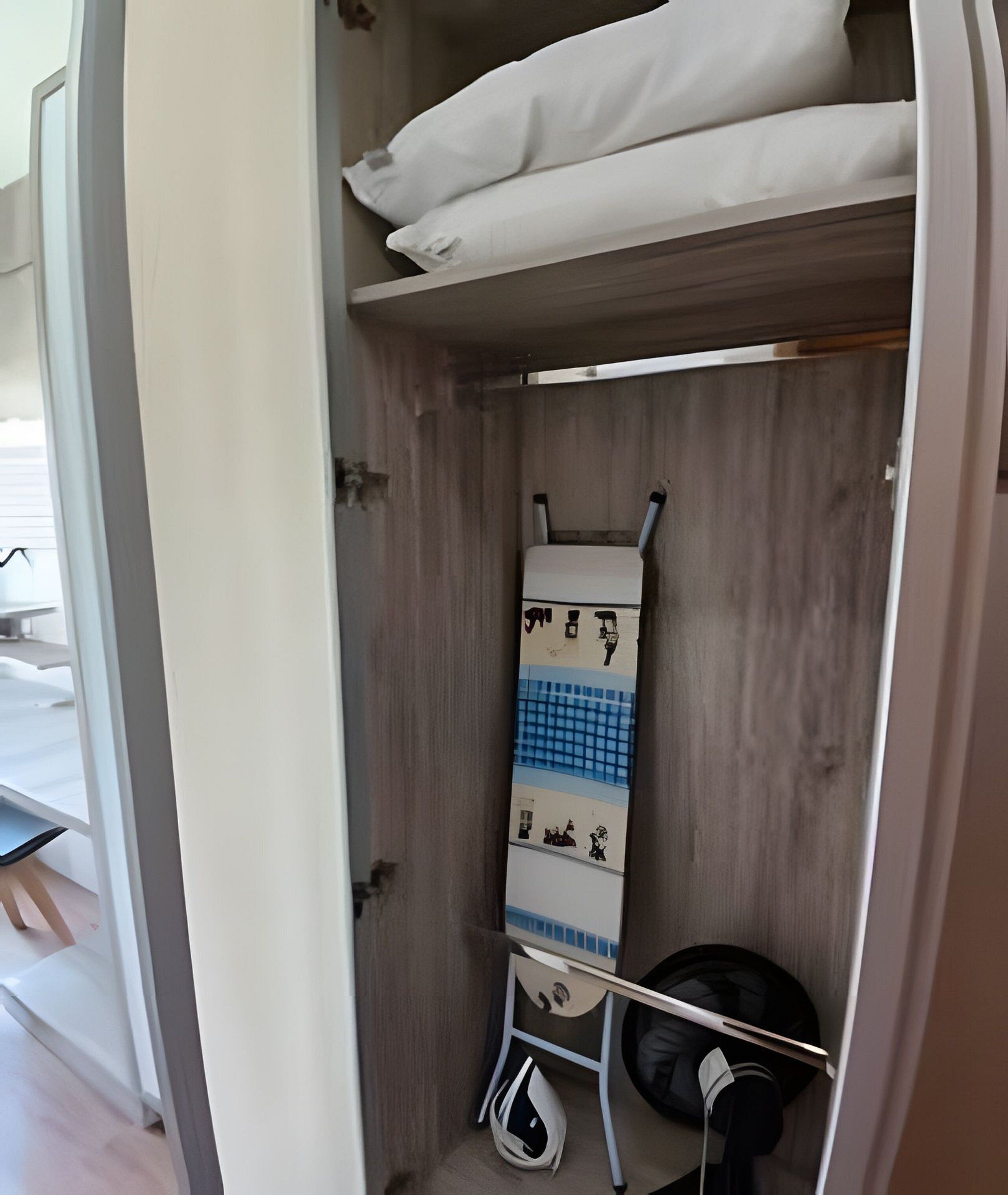 A compact, walk-in closet with a wooden interior, featuring a hanging rack with folded clothes and a small shelf with a white pillow.