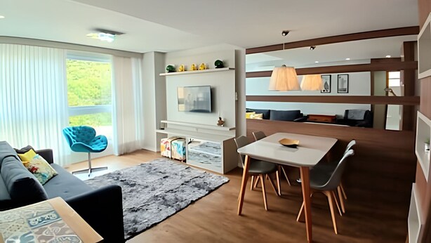 A modern living room with a sofa, a dining table, and a TV. There is a large window with curtains, a rug, and a wooden floor. - Siga Gramado -Ap Lindíssimo 5 min Centro (Gramado)