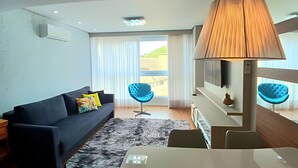 A modern hotel room with a dark-colored sofa, a small round table, a lamp, and a view of a balcony with blue chairs. - Siga Gramado -Ap Lindíssimo 5 min Centro (Gramado)