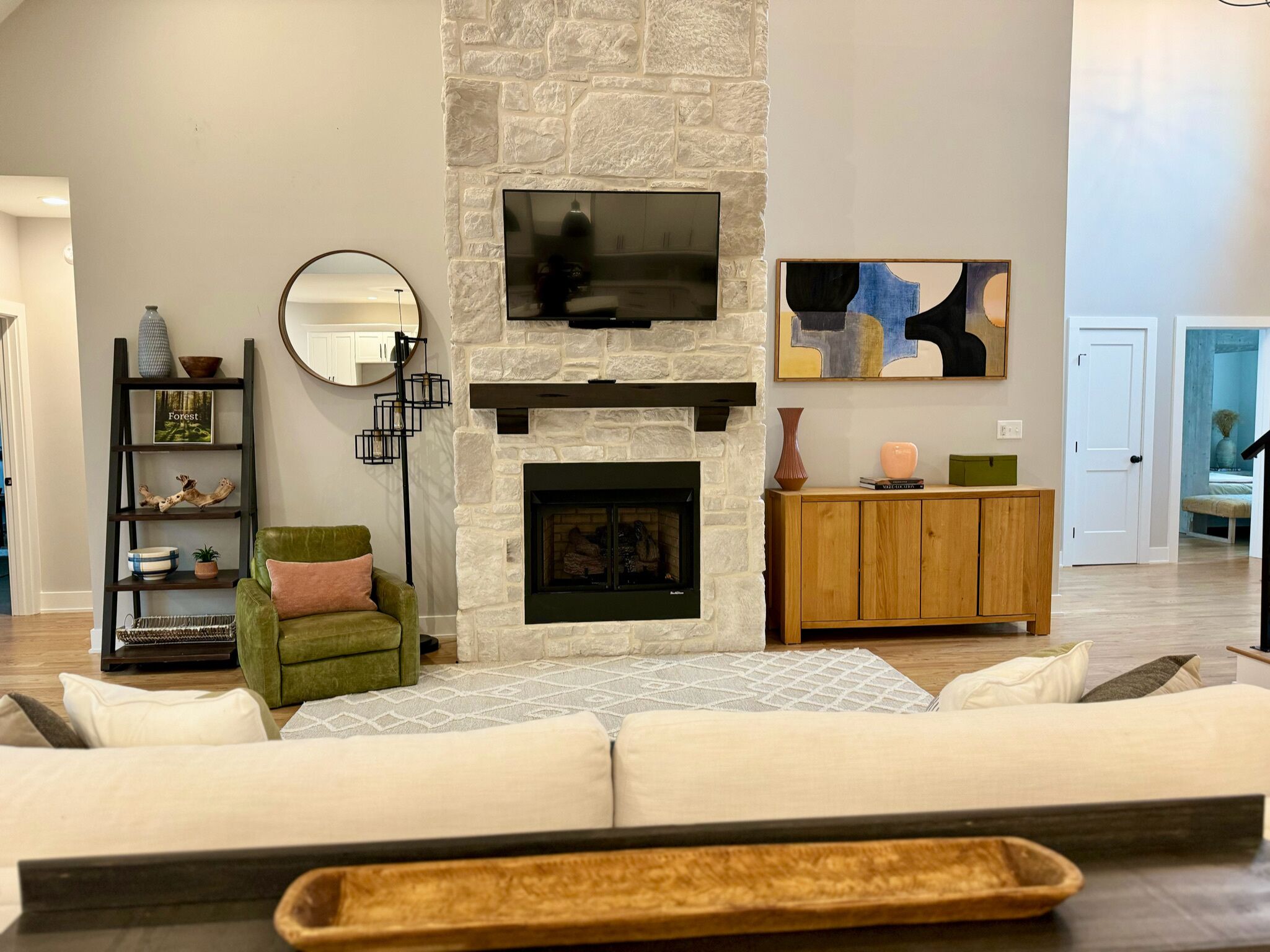 A living room with a stone fireplace, a green armchair, a sofa, a wooden cabinet, and a flat-screen TV mounted above the fireplace.