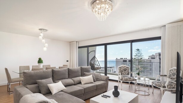 Un salon moderne avec un canapé d’angle gris, une table basse blanche et une grande fenêtre donnant sur la ville.