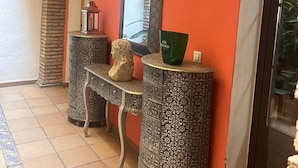 A hallway with a tiled floor, a stone pillar, a mirror, and two ornate side tables with a bust sculpture. - Hostal Lojo (Conil de la Frontera)