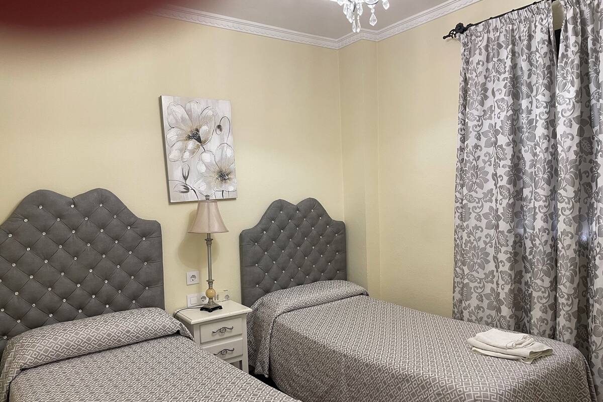 Habitación de hotel con dos camas, una mesita de noche, una lámpara, un cuadro floral y una cortina.