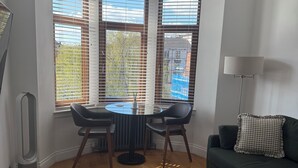 Et hjørneværelse med spisebord, stole, sofa, lampe og store vinduer med skodder.
