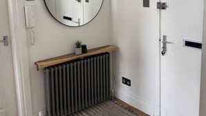 Et værelse med en radiator, et rundt spejl, en træhylde med en plante, en dør med håndtag og en lyskontakt.