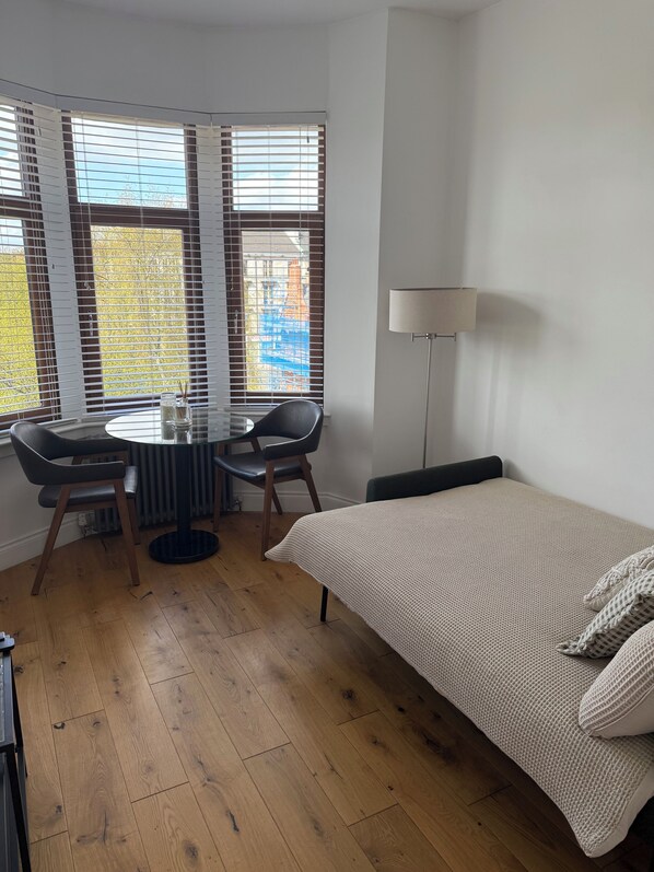 A room with a bed, a round table with chairs, a lamp, and a window with blinds.