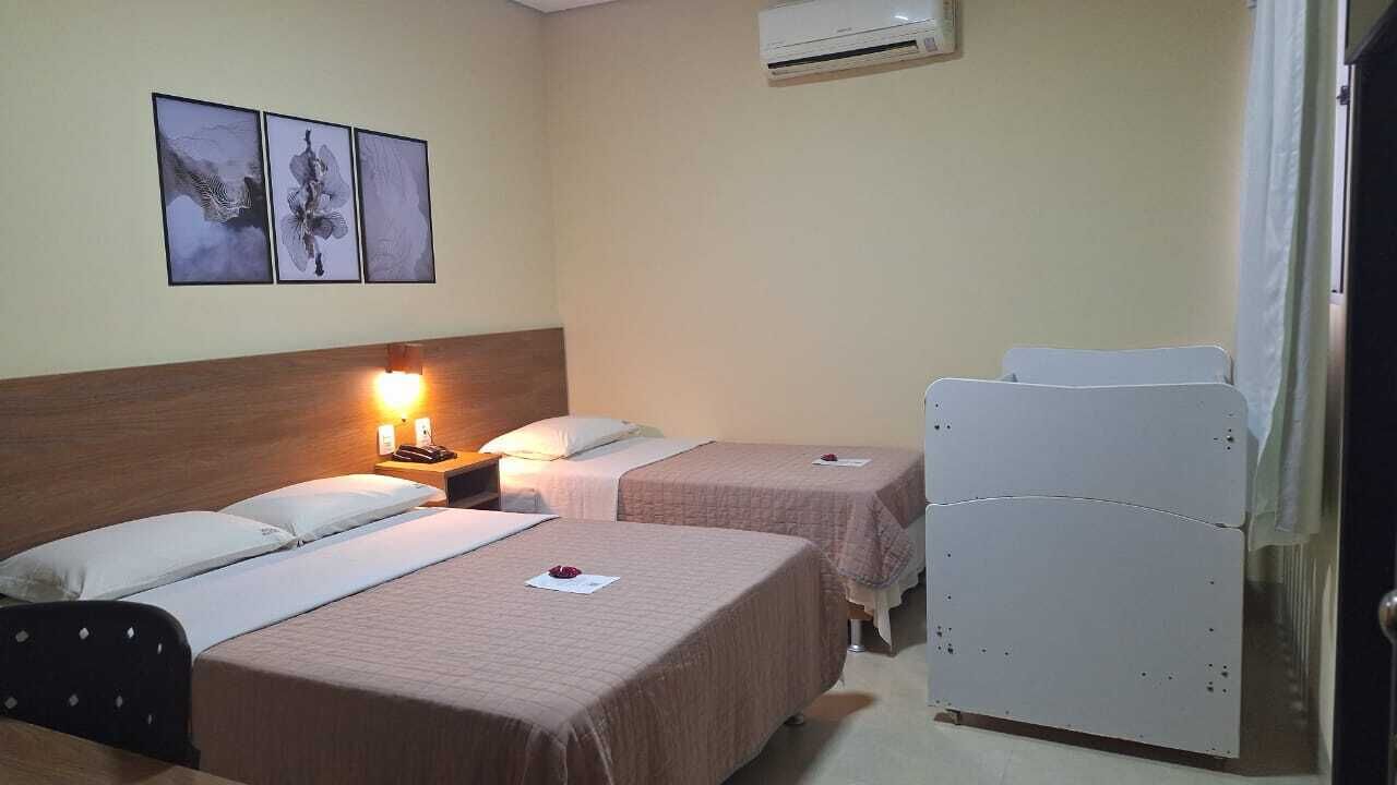 A hotel room with two beds, a bedside table with a lamp, and a wall-mounted air conditioner.