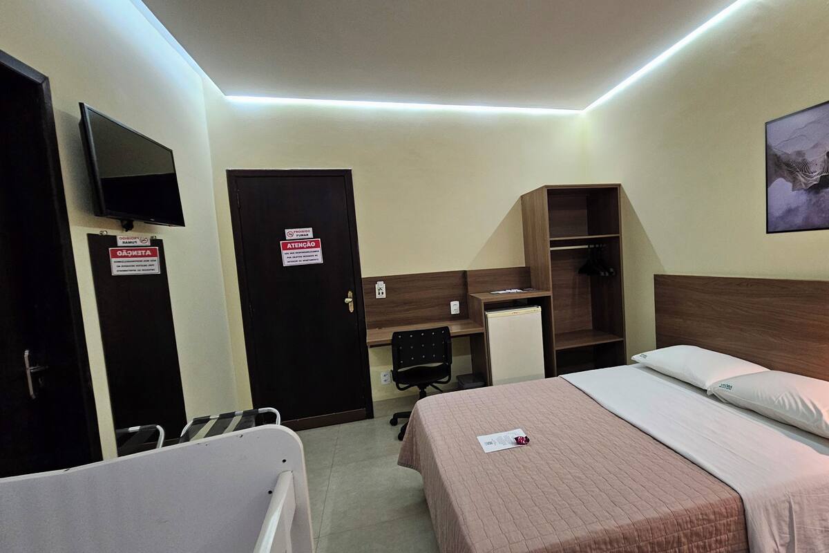 A hotel room with a bed, bedside table, desk, chair, wardrobe, and a television on the wall.