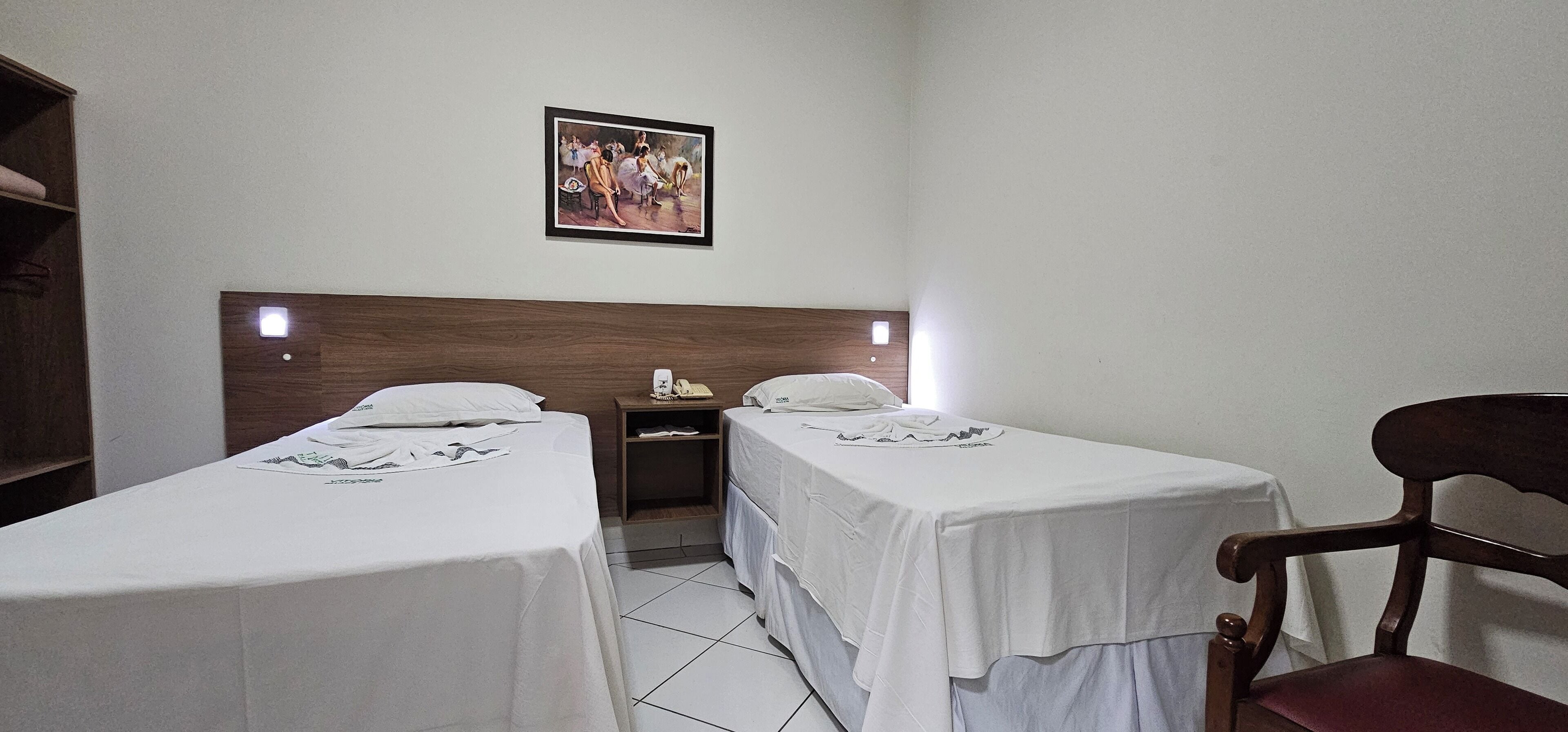 Habitación de hotel con dos camas, cabecera de madera, mesita de noche, una silla y un cuadro en la pared.