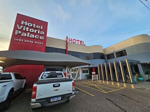 A hotel entrance with a sign for Hotel Vitoria Palace, a white pickup truck parked in front, and a paved area with yellow lines. - VITORIA PALACE HOTEL (Sinop)