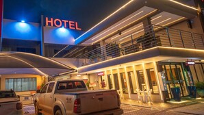 Un hôtel moderne, reconnaissable à son grand panneau lumineux indiquant « HÔTEL », doté d’un espace extérieur couvert et d’un parking où est stationné un camion.