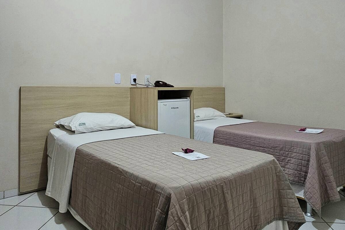 A hotel room with two beds, a small refrigerator, a bedside table, and a ceiling light.