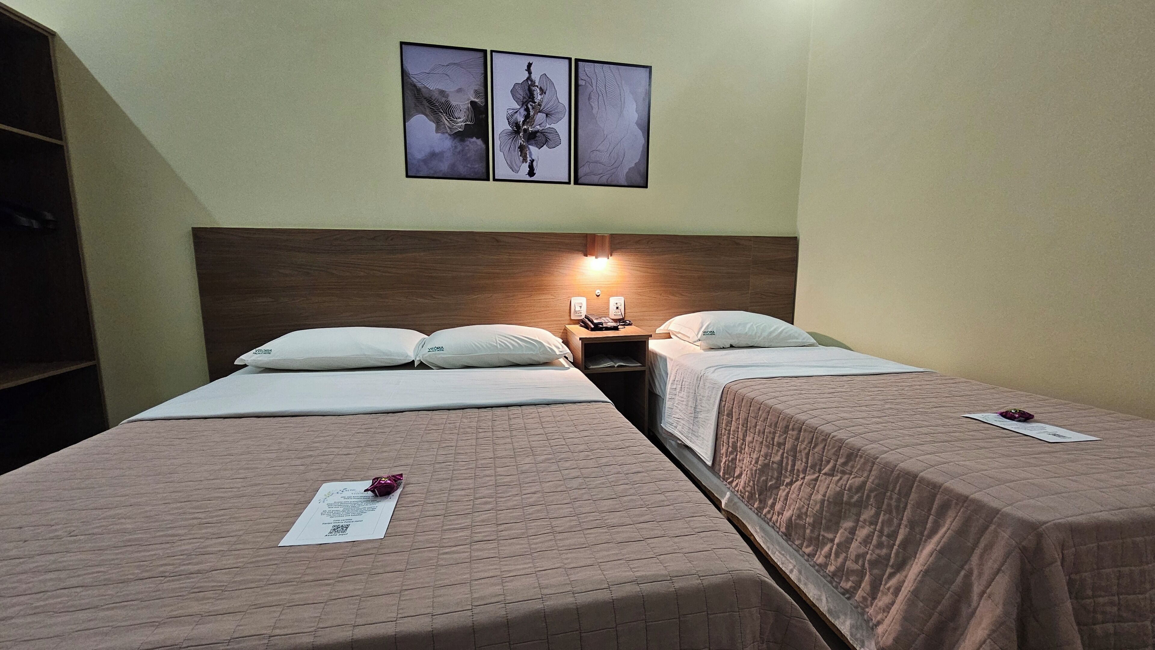 A hotel room with two beds, a wooden headboard, a nightstand with a lamp, and three framed artworks on the wall.