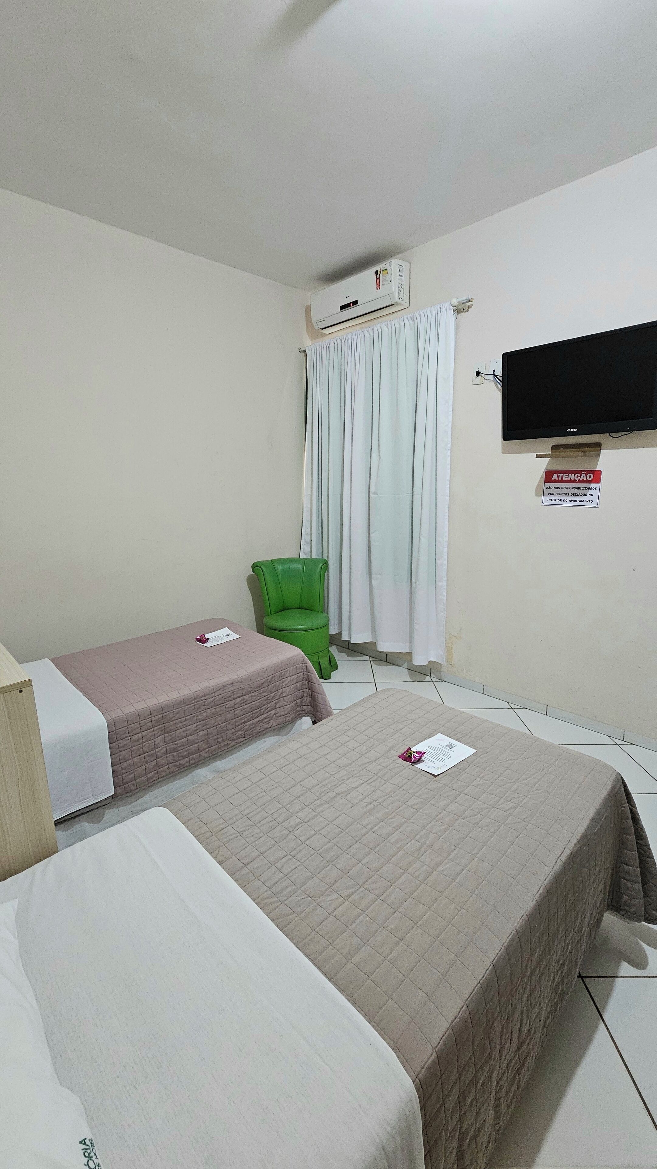 A hotel room with two beds, a green chair, a television, and an air conditioning unit.