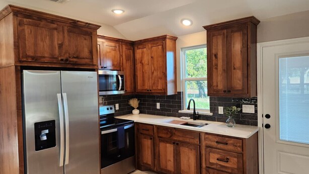 A kitchen with wooden cabinets, stainless steel appliances, a black tiled backsplash, and a window providing natural light. - Country Cabin Getaway (Lometa)